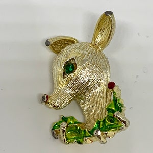 May include: Gold-toned reindeer brooch with a green holly wreath and red berries. The reindeer has a green rhinestone eye.