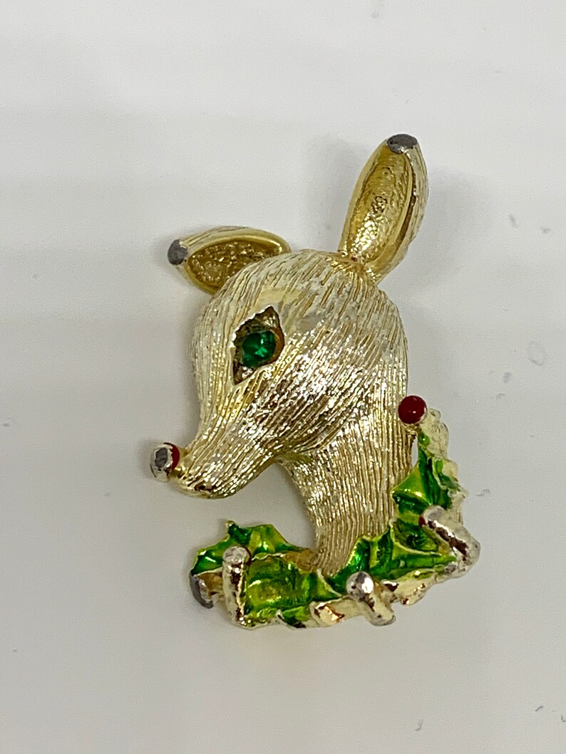 May include: Gold-toned reindeer brooch with a green holly wreath and red berries. The reindeer has a green rhinestone eye.