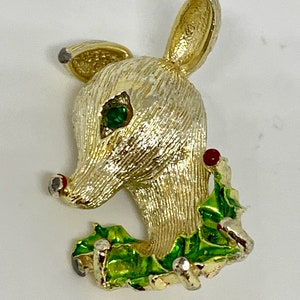 May include: Gold-tone reindeer brooch with a green holly leaf and red berry accents. The reindeer has a green rhinestone eye.