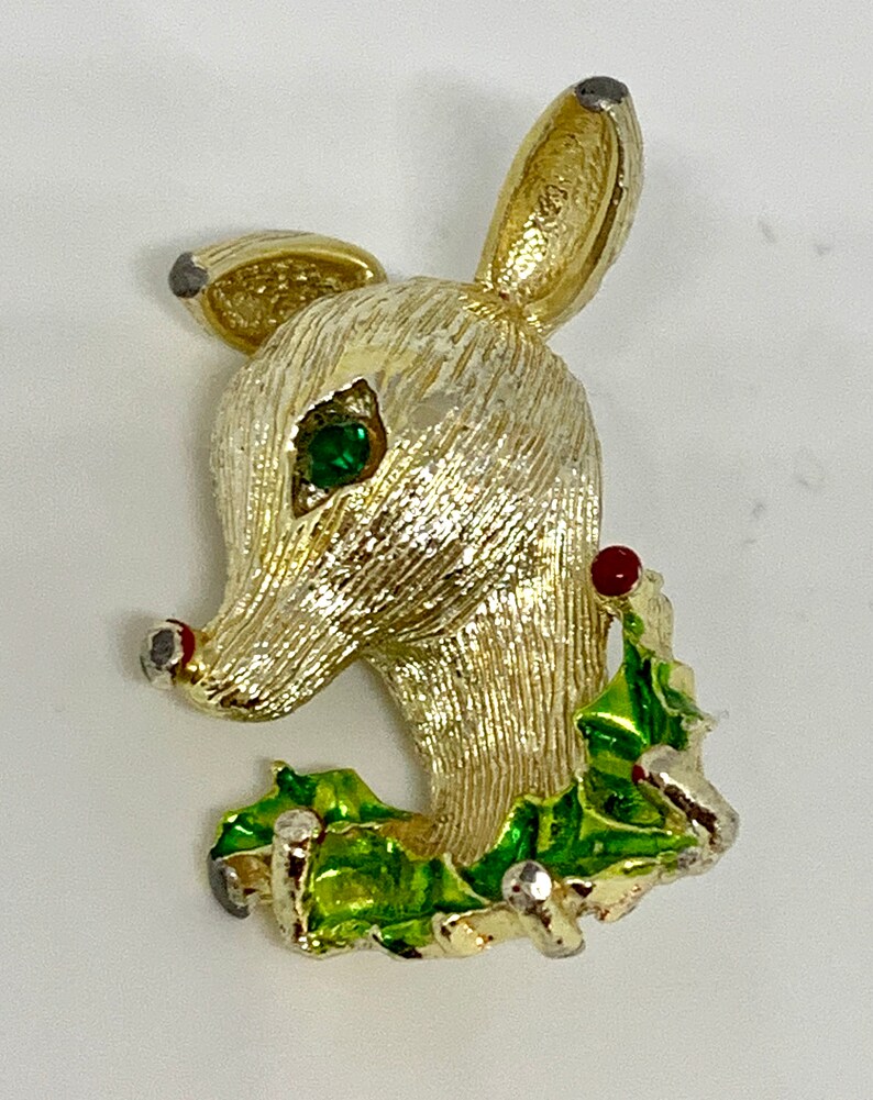 May include: Gold-tone reindeer brooch with a green holly leaf and red berry accents. The reindeer has a green rhinestone eye.