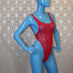 May include: A blue mannequin wearing a red, high-cut swimsuit with thin straps. The mannequin is posed with hands on hips. The background is a geometric pattern in silver and beige.
