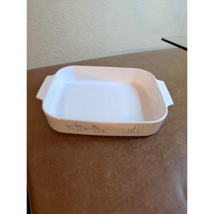 Op de afbeelding: Een vierkante witte keramische ovenschaal met geïntegreerde handgrepen aan de zijkanten. De buitenkant is voorzien van een subtiel, grijs botanisch patroon van dunne grassen en kleine bloemen langs de onderrand, een functionele keukenschaal.