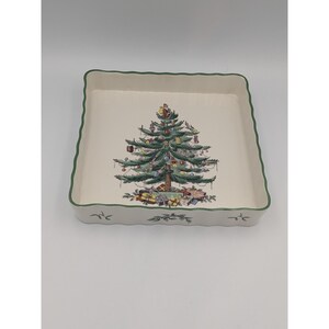 May include: A square ceramic baking dish with a cream-colored interior and a green scalloped rim. The center features a detailed illustration of a decorated Christmas tree with wrapped gifts at the base. The exterior sides feature holly.