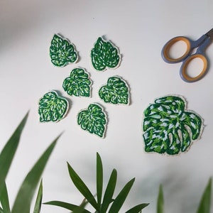 May include: A set of seven green and white embroidered fabric patches shaped like monstera leaves. The patches are arranged on a white surface with a pair of scissors in the top right corner.