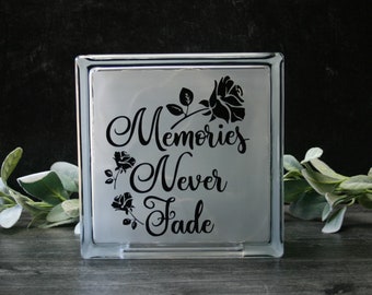 May include: A clear glass block featuring the words "Memories Never Fade" in a stylish script, with black rose motifs. The block is designed to stand upright and is suitable for decorative purposes.
