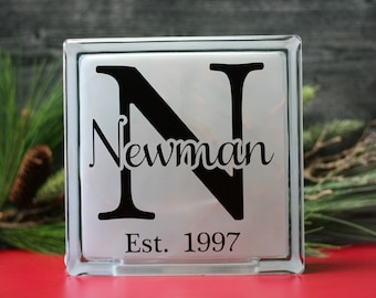 May include: A white glass block with a black letter N and the name "Newman" written in cursive below it. The text "Est. 1997" is written below the name.