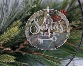 May include: A clear, circular Christmas ornament engraved with the words "Santa's Magic Key." A small silver-coloured charm of Santa Claus and a key hangs from the top. The ornament is set against a backdrop of pine branches.