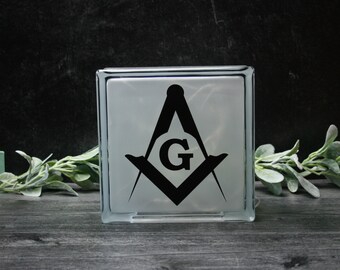 May include: A white glass block with a black and white image of a compass and square with a "G" in the centre. The block is sitting on a wooden surface.
