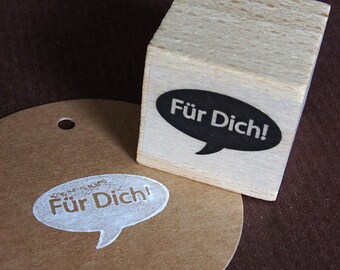 May include: A wooden block stamp with a black speech bubble that reads "Für Dich!"  The stamp is used to create a white speech bubble with the same text on a brown paper surface.