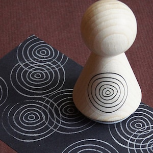 Könnte beinhalten: Ein Stempel aus Holz mit einem konzentrischen Kreisdesign. Der Stempel wird verwendet, um ein sich wiederholendes Muster von Kreisen auf einer schwarzen Oberfläche zu erstellen.