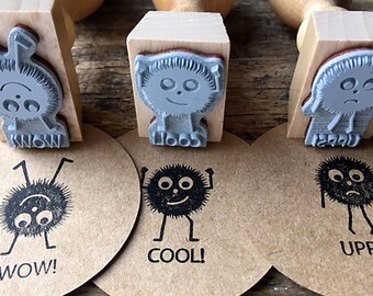 May include: Three wooden stamps with cute fuzzy monster designs. The stamps are on a wooden surface with three round brown paper circles with the words "WOW!" "COOL!" and "UPP!" printed on them. The stamps are gray and the designs are black.