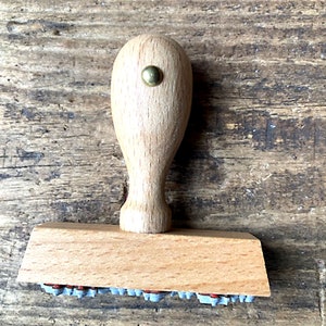 May include: A wooden stamp with a light brown handle and a rectangular base. The base has a row of small, gray rubber stamps.