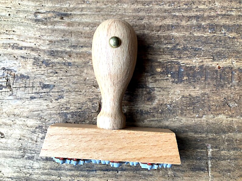 May include: A wooden stamp with a light brown handle and a rectangular base. The base has a row of small, gray rubber stamps.