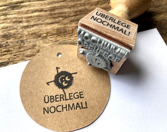 May include: A wooden stamp with the text "Ueberlege nochmal!" on it. The stamp is being used to create a custom label with the same text on a brown paper circle.