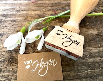 May include: A wooden stamp with the words "von Herzen" and a heart symbol, alongside a stamped image on a small brown card. Two white snowdrop flowers with green stems are also present.