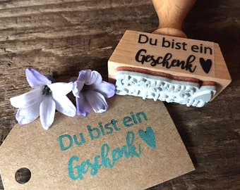 May include: A wooden stamp with the text "Du bist ein Geschenk" and a small black heart. The stamp is on a wooden surface with a brown paper tag that says "Du bist ein Geschenk" in teal with a teal heart.