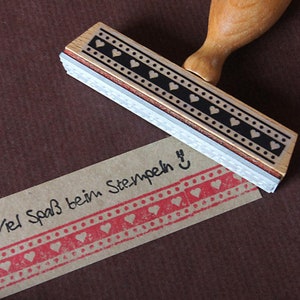 Könnte beinhalten: Ein Holzstempel mit einem schwarz-weißen Herzmuster. Der Stempel wird verwendet, um ein rotes Herzmuster auf einem braunen Papieranhänger zu erstellen. Der Anhänger lautet "Viel Spaß beim Stempeln!"