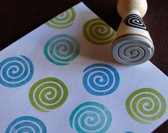 May include: A white card with a repeating pattern of blue and green spiral designs. A wooden stamp with a gray spiral design is on top of the card.