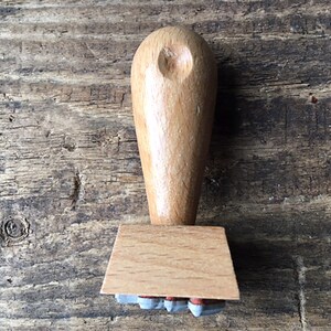 K&ouml;nnte beinhalten: Ein Stempel aus Holz mit einem hellbraunen Griff und einem rechteckigen Sockel. Der Sockel hat eine Reihe kleiner, grauer Gummistempel.