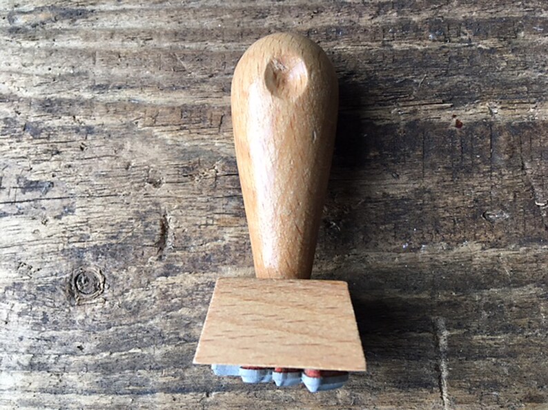 K&ouml;nnte beinhalten: Ein Stempel aus Holz mit einem hellbraunen Griff und einem rechteckigen Sockel. Der Sockel hat eine Reihe kleiner, grauer Gummistempel.
