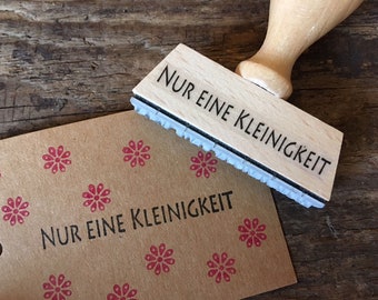 May include: Wooden stamp with the text "NUR EINE KLEINIGKEIT" in black lettering. The stamp is on a brown paper background with a repeating pattern of red flowers.