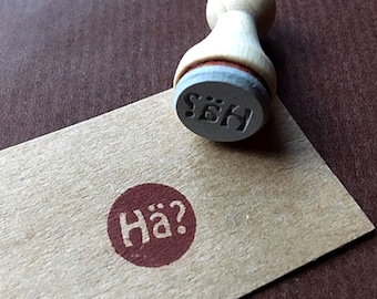 May include: A brown wooden stamp with the text "HÄ?" in a white circle on a brown paper tag. The stamp is also on a brown surface.