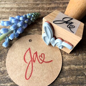 K&ouml;nnte beinhalten: Ein Stempel aus Holz mit einem kursiv geschriebenen "Jae" und einem Herzdesign. Der Stempel wird verwendet, um ein rotes "Jae" mit einem Herz auf einem braunen Papieretikett zu erstellen.