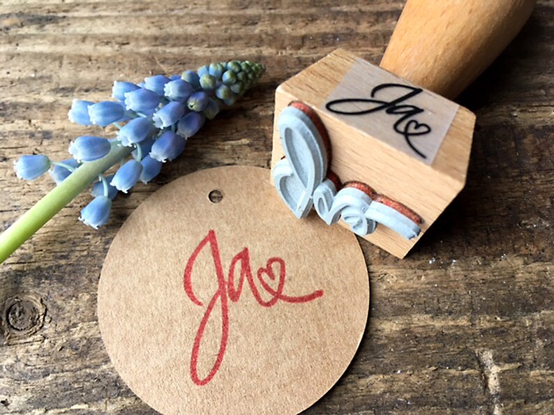 K&ouml;nnte beinhalten: Ein Stempel aus Holz mit einem kursiv geschriebenen "Jae" und einem Herzdesign. Der Stempel wird verwendet, um ein rotes "Jae" mit einem Herz auf einem braunen Papieretikett zu erstellen.