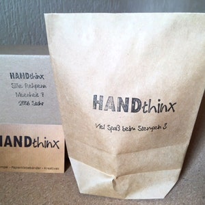 May include: A brown paper bag with the text "HANDthinx" and the phrase "Viel Spa&szlig; beim Stempeh" printed on it. The bag is sitting on a wooden surface next to a small brown box with the text "HANDthinx" and contact information printed on it.