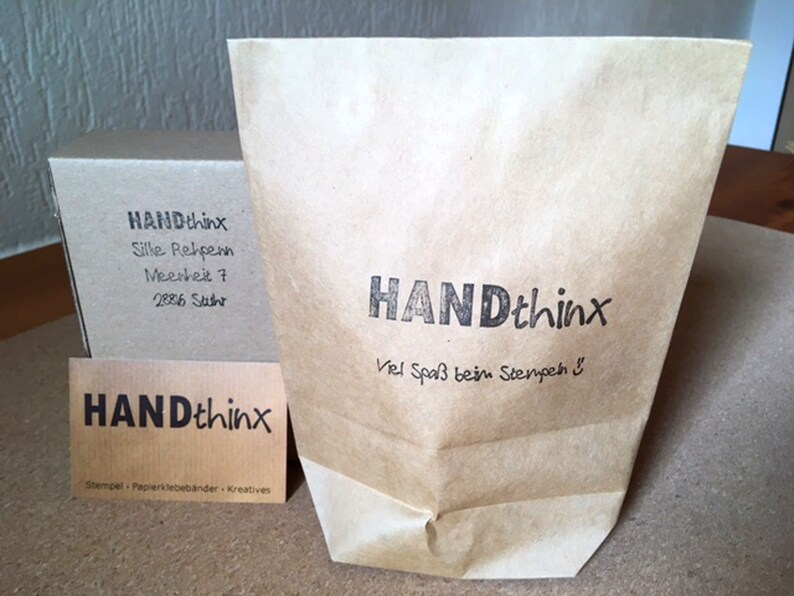 May include: A brown paper bag with the text "HANDthinx" and the phrase "Viel Spa&szlig; beim Stempeh" printed on it. The bag is sitting on a wooden surface next to a small brown box with the text "HANDthinx" and contact information printed on it.
