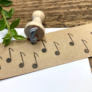 Könnte beinhalten: Ein Stempel aus Holz mit einem Notenmotiv. Der Stempel wird verwendet, um mehrere Notenabdrücke auf einem braunen Papieretikett zu erstellen.