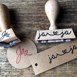 K&ouml;nnte beinhalten: Zwei Holzstempel mit schwarzer Tinte. Der erste Stempel sagt "ja" mit einem Herzsymbol zwischen den Buchstaben. Der zweite Stempel sagt "ja" mit einem Herzsymbol zwischen den Buchstaben. Die Stempel befinden sich auf einer Holzoberfl&auml;che mit zwei braunen Papieretiketten, die den gleichen Text wie die Stempel haben.