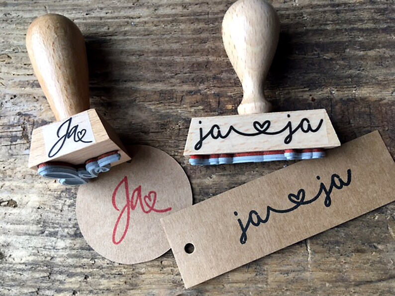 K&ouml;nnte beinhalten: Zwei Holzstempel mit schwarzer Tinte. Der erste Stempel sagt "ja" mit einem Herzsymbol zwischen den Buchstaben. Der zweite Stempel sagt "ja" mit einem Herzsymbol zwischen den Buchstaben. Die Stempel befinden sich auf einer Holzoberfl&auml;che mit zwei braunen Papieretiketten, die den gleichen Text wie die Stempel haben.