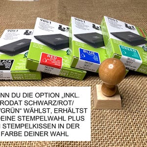 May include: Four boxes of Trodat stamp pads in black, red, blue, and green. The boxes are labeled with the color of the ink pad. A wooden stamp is in the foreground. The text on the boxes reads "INKL. TRODAT SCHWARZ/ROT/BLAU/GR&Uuml;N" and "WENN DU DIE OPTION "INKL. TRODAT SCHWARZ/ROT/BLAU/GR&Uuml;N" W&Auml;HLST, ERH&Auml;LTST DU DEINE STEMPELWAHL PLUS EIN STEMPELKISSEN IN DER FARBE DEINER WAHL".