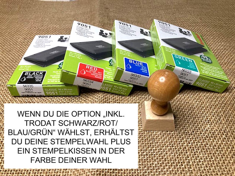 May include: Four boxes of Trodat stamp pads in black, red, blue, and green. The boxes are labeled with the color of the ink pad. A wooden stamp is in the foreground. The text on the boxes reads "INKL. TRODAT SCHWARZ/ROT/BLAU/GR&Uuml;N" and "WENN DU DIE OPTION "INKL. TRODAT SCHWARZ/ROT/BLAU/GR&Uuml;N" W&Auml;HLST, ERH&Auml;LTST DU DEINE STEMPELWAHL PLUS EIN STEMPELKISSEN IN DER FARBE DEINER WAHL".