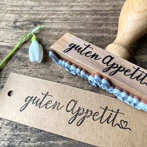 May include: Wooden stamp with the text "guten Appetit" in black script with a heart at the end. The stamp is on a brown paper tag with the same text stamped in black.