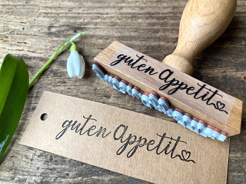 May include: Wooden stamp with the text "guten Appetit" in black script with a heart at the end. The stamp is on a brown paper tag with the same text stamped in black.