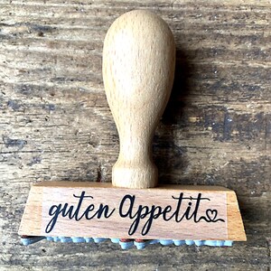 May include: Wooden stamp with a brown handle and a rectangular base. The base has the text "guten Appetit" with a small heart symbol.
