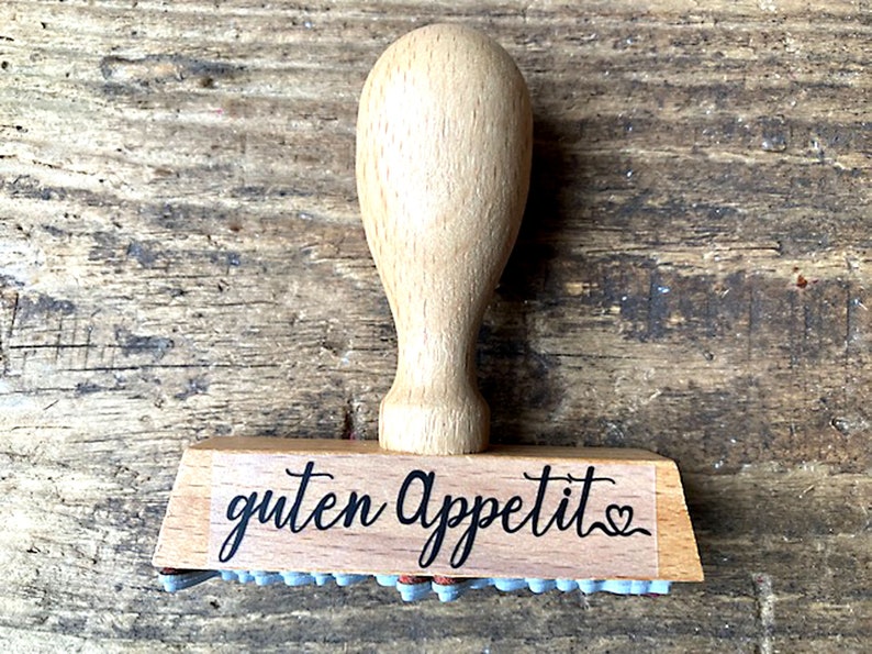 May include: Wooden stamp with a brown handle and a rectangular base. The base has the text "guten Appetit" with a small heart symbol.