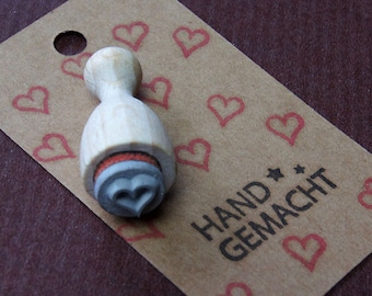 May include: A wooden stamp with a heart-shaped design, next to a brown tag with red heart illustrations and the words "HAND GEMACHT". The stamp has a wooden handle and a gray rubber stamp head. The tag has a hole at the top.
