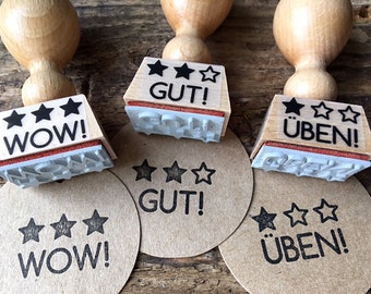 May include: Three wooden stamps with black ink pads. Each stamp has three stars and a word printed on it. The words are "WOW!" "GUT!" and "ÜBEN!" The stamps are sitting on brown paper circles with the same words printed on them.