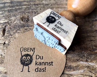 May include: A wooden stamp with a black and white illustration of a fuzzy creature with the text "ÜBEN! Du kannst das!"  The stamp is being used to create an impression on a brown paper tag.