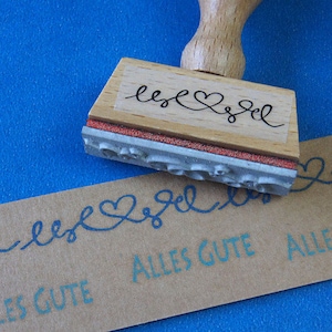 Könnte beinhalten: Ein Stempel aus Holz mit einer durchsichtigen Acryloberseite. Der Stempel trägt die Aufschrift "lesel" mit einem Herzsymbol in der Mitte. Der Stempel wird verwendet, um eine Linie von "lesel" mit Herzen auf einem braunen Papieranhänger zu erstellen. Der Anhänger trägt auch den Text "Alles Gute" aufgedruckt.