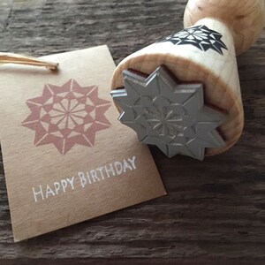 Könnte beinhalten: Ein Holzstempel mit einem Sternmuster und einem passenden Anhänger mit der Aufschrift "HAPPY BIRTHDAY". Der Stempel hat einen Holzgriff und ein detailliertes Sternmuster. Der Anhänger ist braun mit einem rosa Sternmuster.