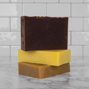 May include: Three bars of soap stacked on top of each other. The top bar is dark brown, the middle bar is yellow, and the bottom bar is light brown.