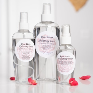 May include: Three clear glass bottles with spray tops filled with rose water hydrating toner. The bottles are labeled with a white circle and black text that reads "Rose Water Hydrating Toner" and "Distilled water & Rose".