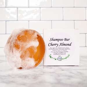May include: A round, amber-colored shampoo bar with foamy lather, next to a white box. The box has a label that reads "Shampoo Bar Cherry Almond" with a list of ingredients and the shop's name.