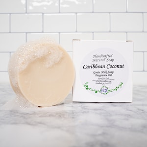 May include: A white box with a green vine design and the text "Handcrafted Natural Soap Caribbean Coconut Goats Milk Soap Fragrance Oil External Use Only etsy.com/shop/MangroveBubbles Handmade in Ararat NC 5 oz"  A round, white soap bar with a white foam on top.