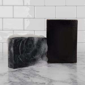 May include: A black bar of soap with white suds, next to a black box. The soap has a textured surface and is rectangular in shape. The box is also black and rectangular. The background is a white tile wall.