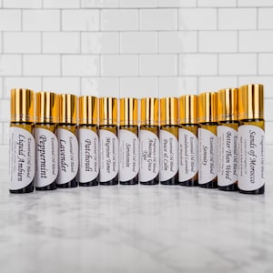 May include: A row of 12 gold and brown essential oil roller bottles with white labels. The labels list the names of the essential oil blends, such as "Liquid Ambien", "Peppermint", "Lavender", "Patchouli", "Migraine Tamer", "Serotonin", "Amazing Grace", "Peace & Calm", "Sandalwood Lavender", "Serenity", "Better Than Weed", and "Sands of Morocco".